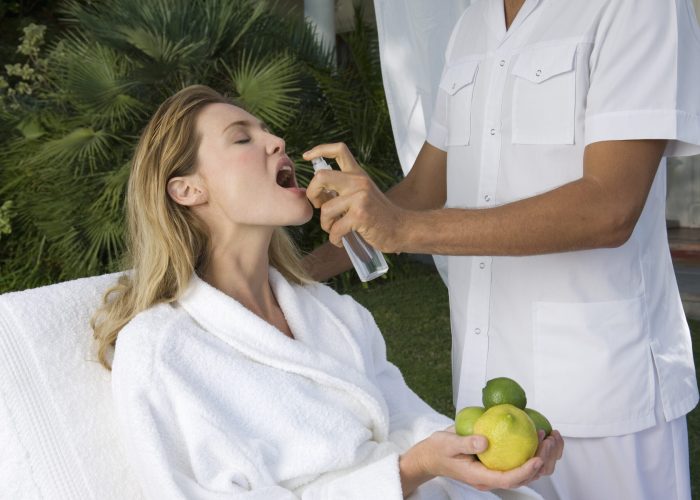 Blond woman wearing white bathrobe sitting outdoors, holding citrus fruit, a beautician spraying water in her mouth.